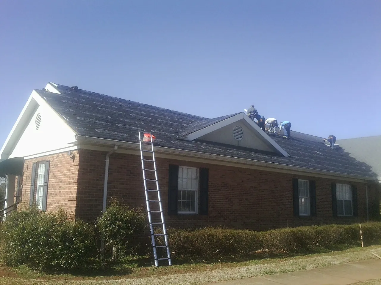 Crew tearing shingles off a brick ranch roof with a ladder set — Emergency Roof Repair work by our crew in Talladega