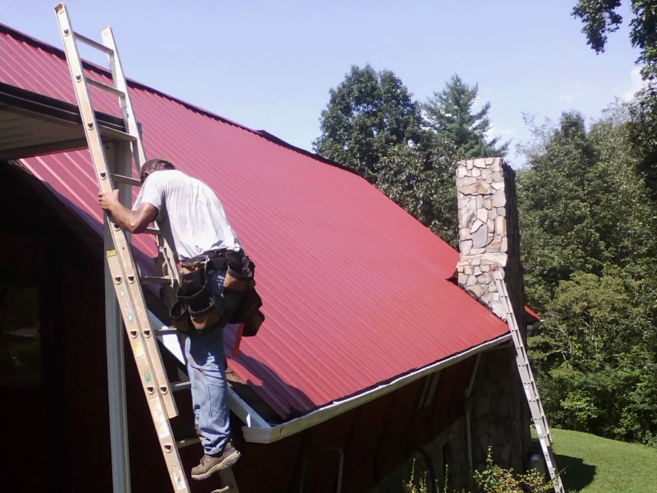 Roofer on a ladder beside a red metal roof and stone chimney — Soffit & Fascia Repair by Roofing Brothers in Talladega